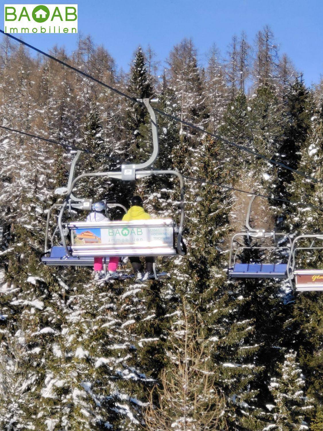 Skifahrer auf einem Sessellift, der über eine winterliche Landschaft mit verschneiten Bäumen fährt.