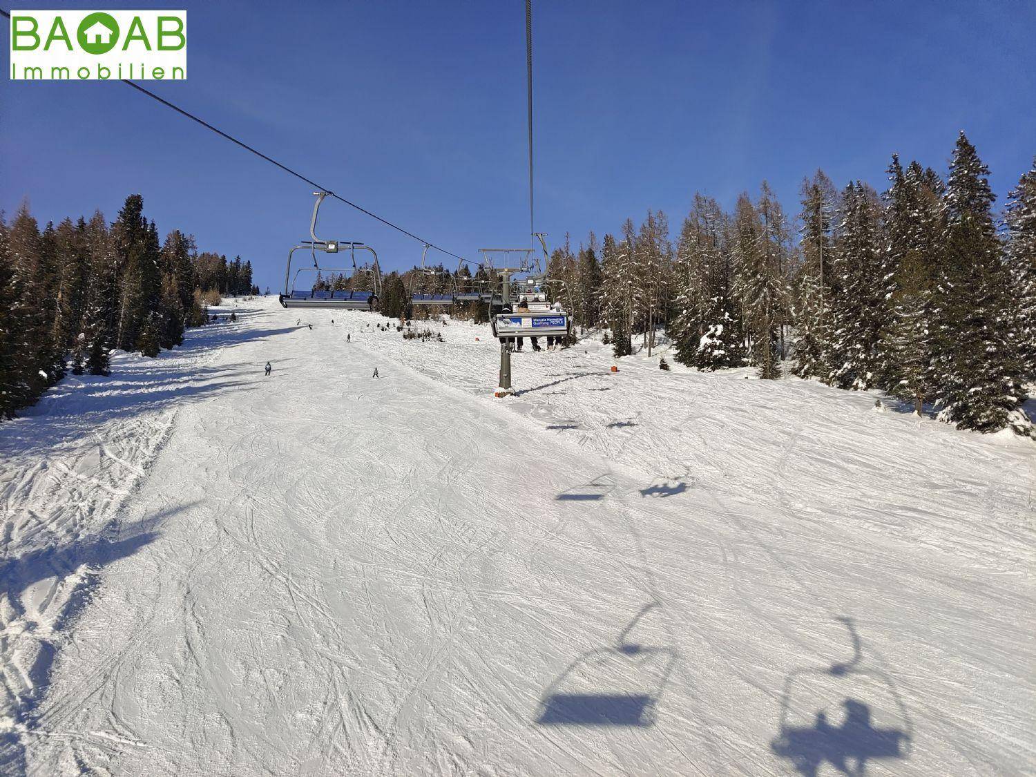 Sonnige Skipiste mit einem Sessellift, Skifahrern und schneebedeckten Bäumen unter blauem Himmel.