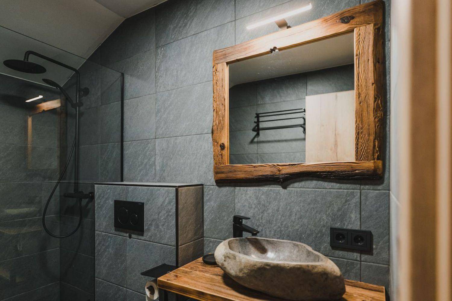 Detailansicht des Badezimmers mit Steinwaschbecken, Holzspiegel und grauen Wandfliesen.