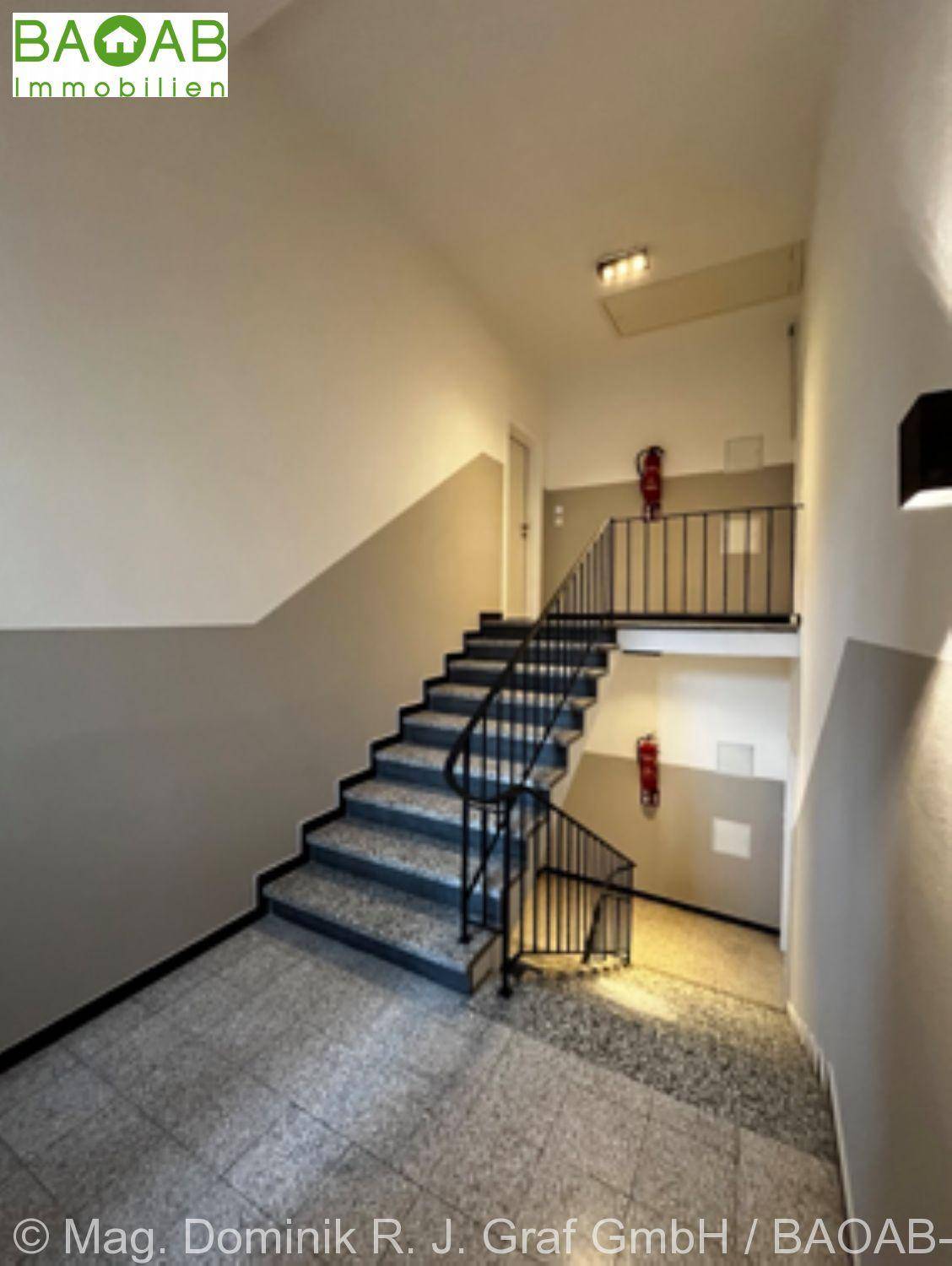 Geräumiges Treppenhaus mit mehreren Absätzen und einem Feuerlöscher an der Wand.