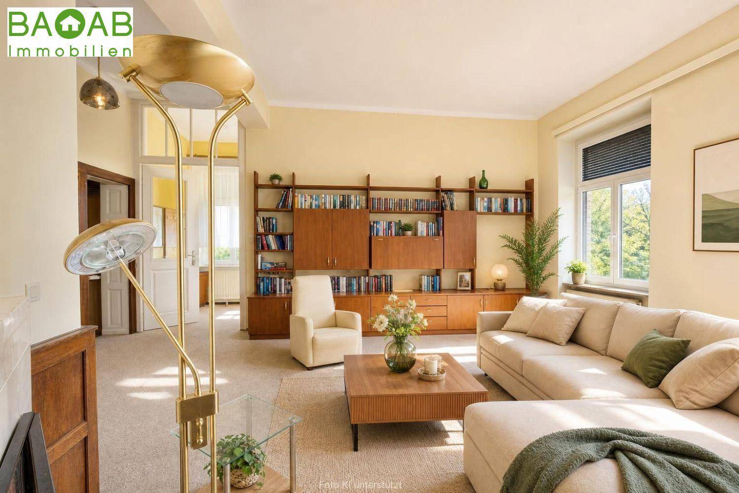 Geräumiges Wohnzimmer mit beigem Sofa, Bücherregal und großem Fenster mit Blick ins Grüne.