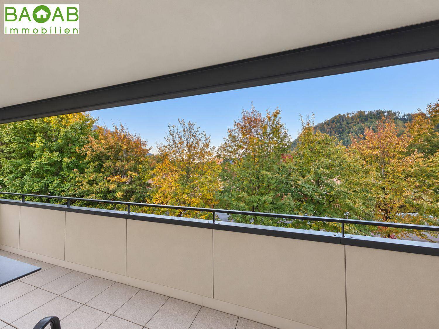 Weitläufiger Blick vom Balkon auf die grünen Bäume und die umliegende Berglandschaft unter blauem Himmel.
