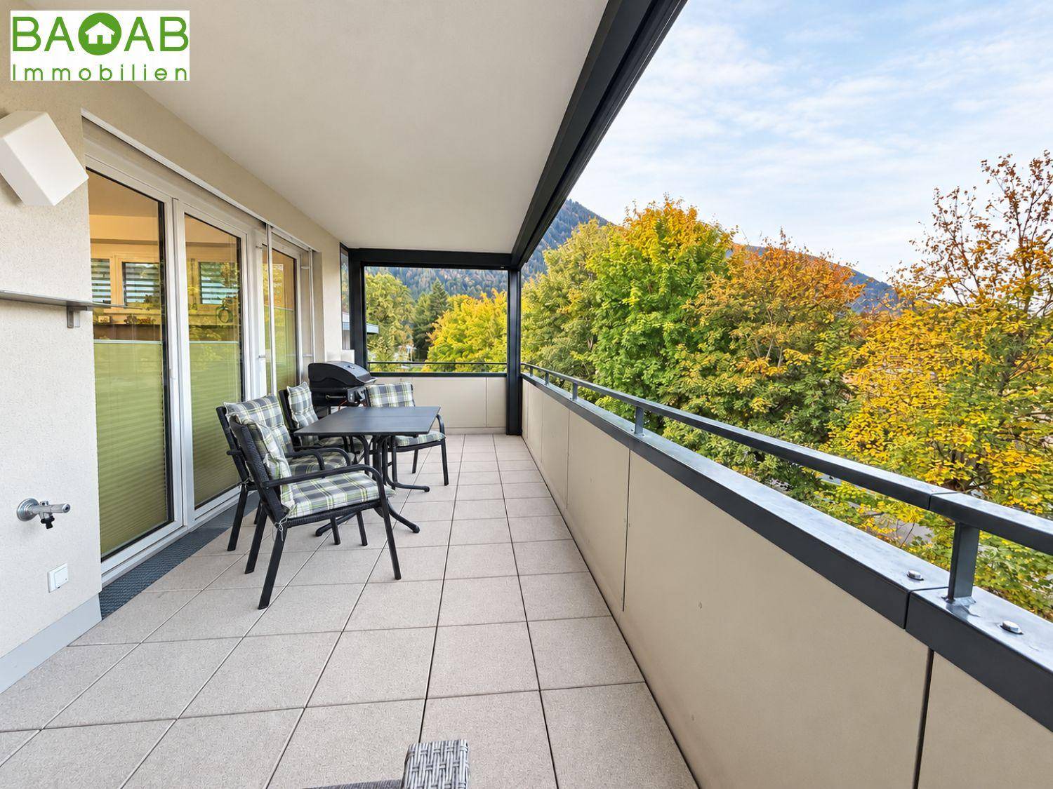 Geräumiger Balkon mit Fliesenboden, Esstisch und Stühlen, umgeben von herbstlichen Bäumen und Bergblick.