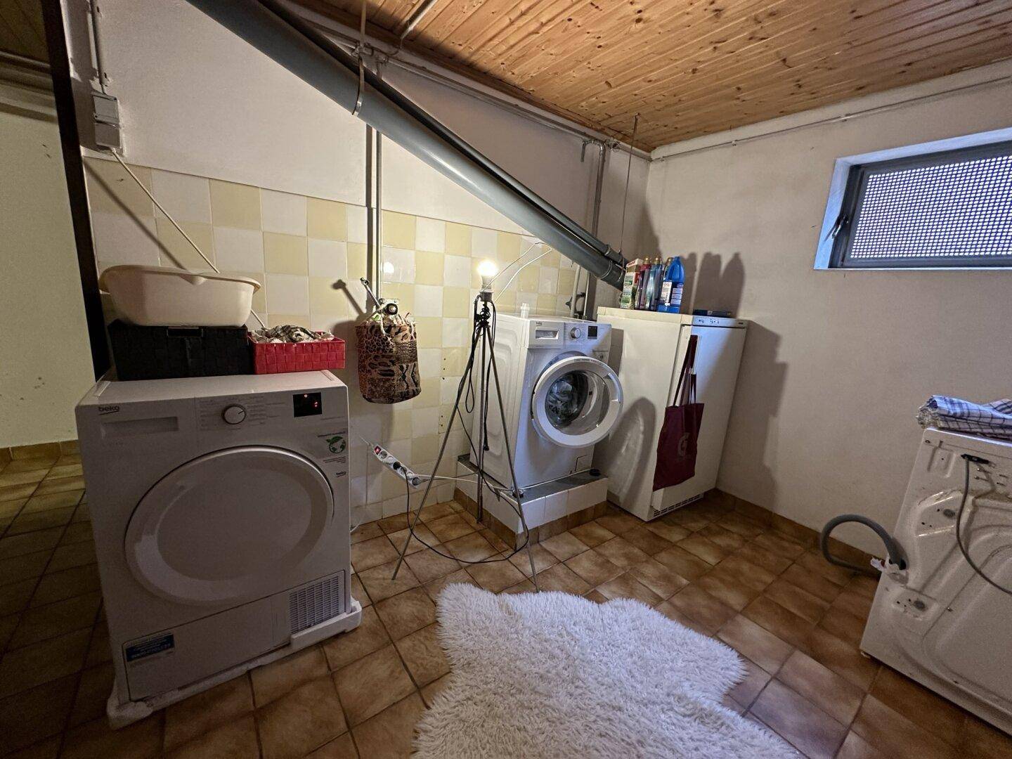 Funktionaler Waschraum mit Waschmaschine, Trockner, gefliesten Wänden und Holzdecke.