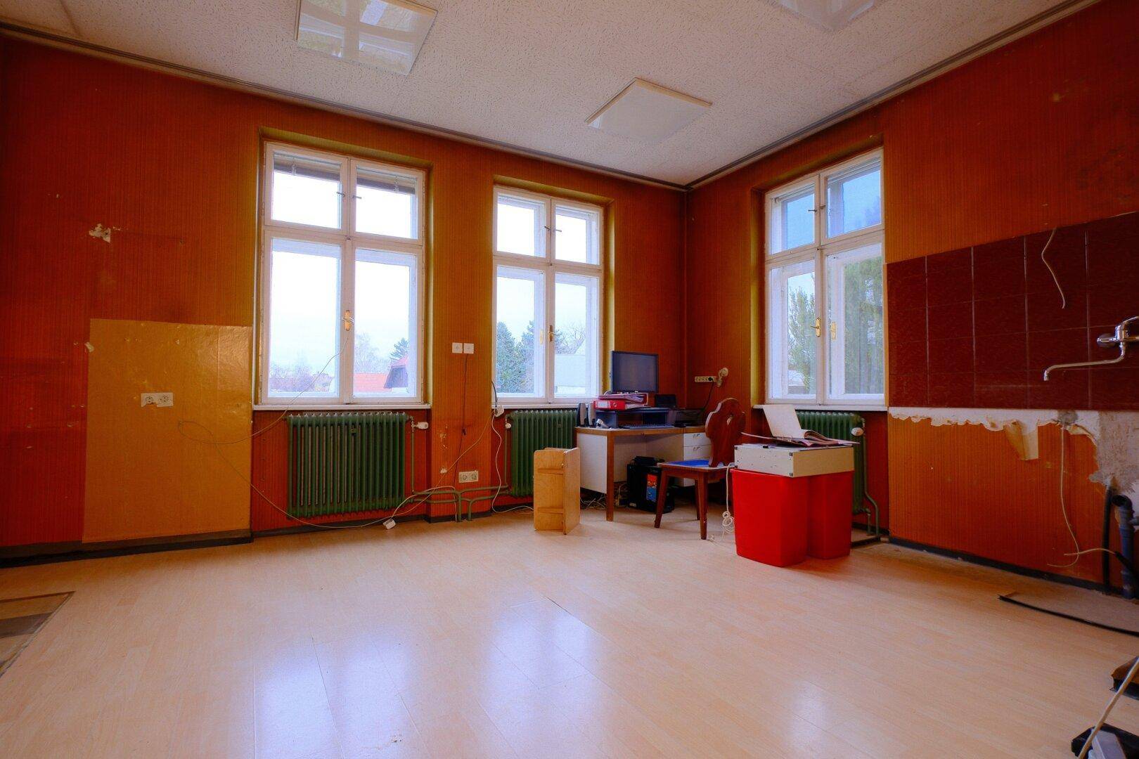 Geräumiger Raum mit orangefarbenen Wänden, Holzfußboden und mehreren Fenstern, renovierungsbedürftig.