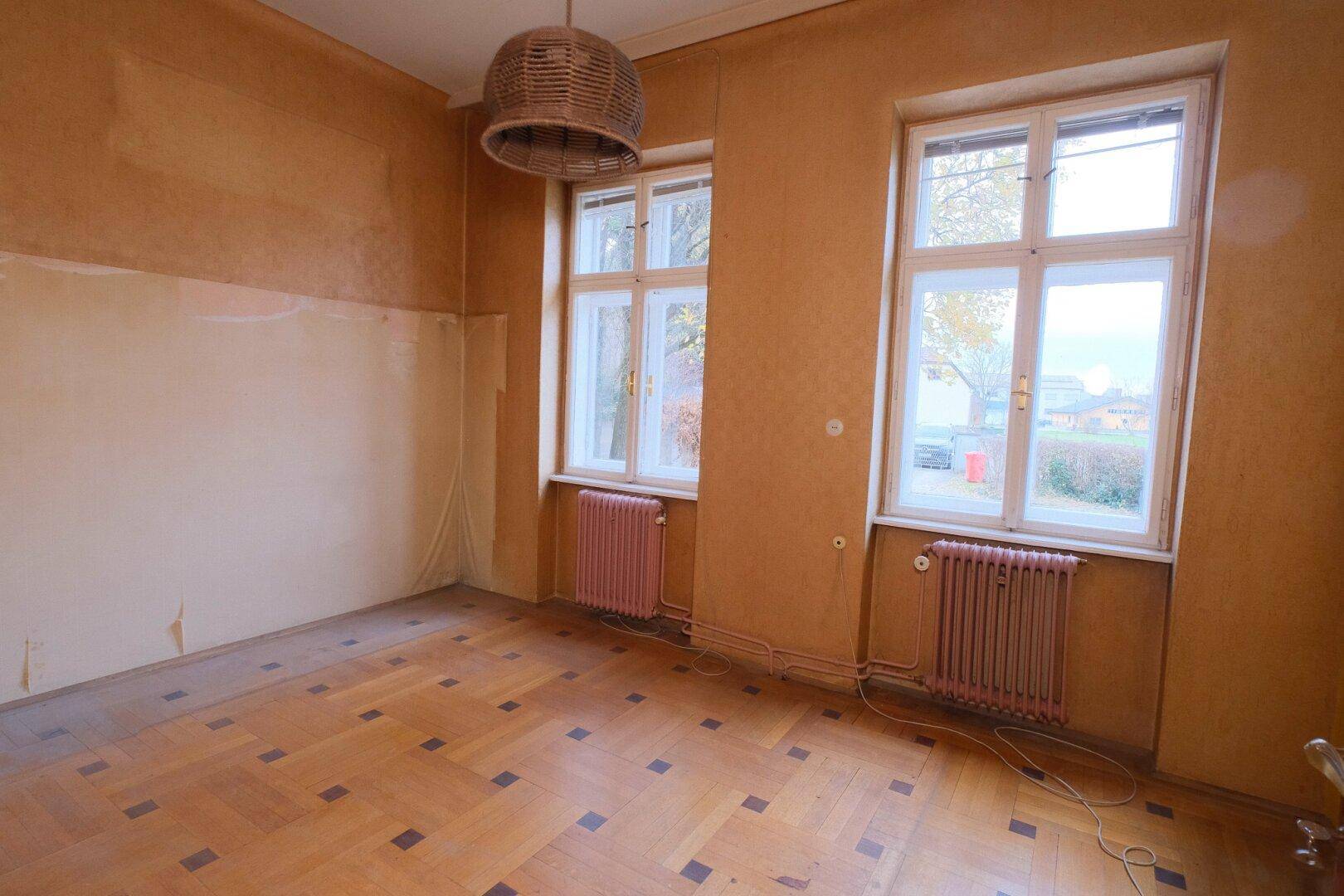 Zimmer mit gemustertem Parkettboden, hellen Wänden und zwei großen Fenstern.