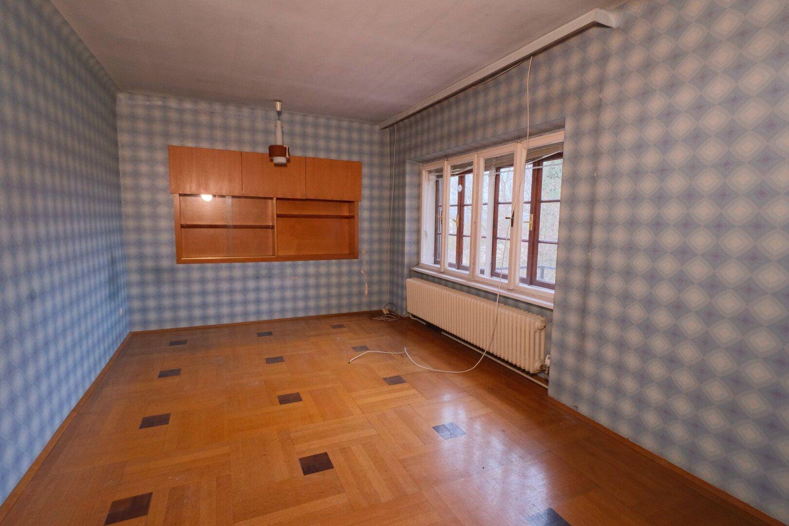 Schlafzimmer mit gemustertem Parkettboden, blauer Tapete und einem Fenster mit Heizkörper.