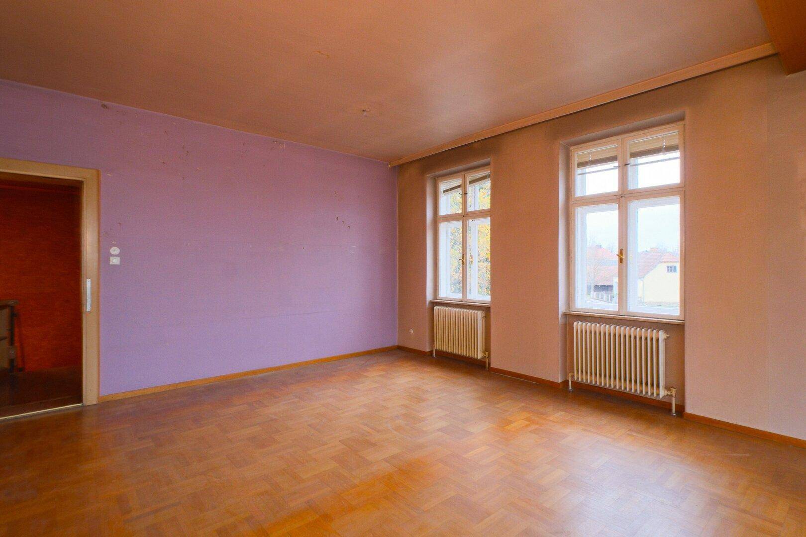 Schlafzimmer mit Parkettboden, einer lila Wand und zwei Fenstern mit Heizkörpern.