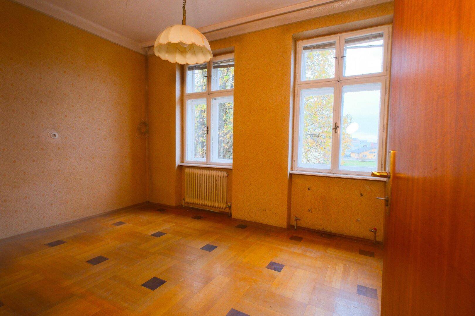 Zimmer mit gemustertem Parkettboden, gelber Tapete und zwei Fenstern mit Heizkörpern.