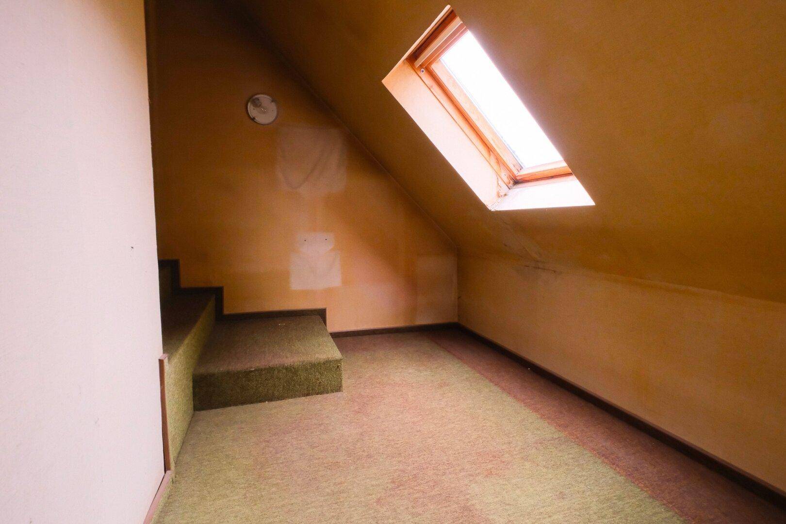 Dachzimmer mit schräger Decke, Teppichboden und einem Dachfenster für natürliches Licht.