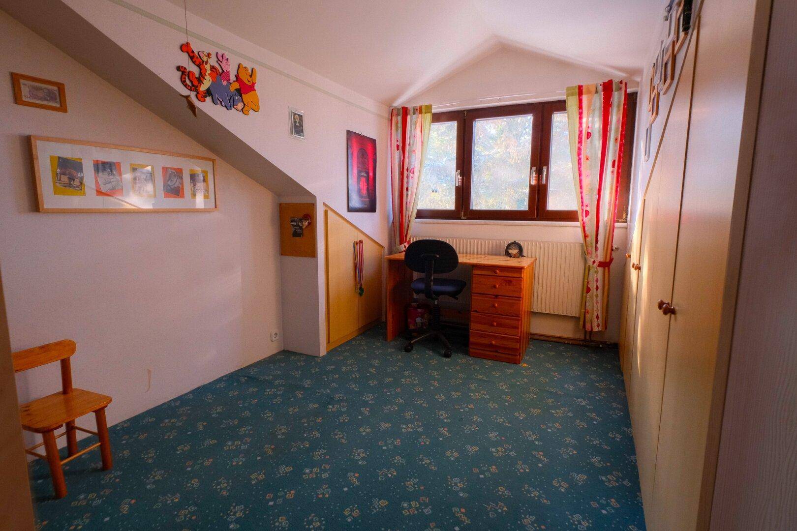 Helles Kinderzimmer im Dachgeschoss mit Schrägen, Teppichboden und einem Schreibtischbereich.
