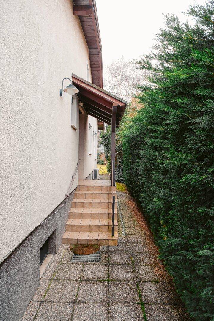 Seitlicher Zugang zum Haus mit gefliestem Weg, Treppe und hoher Hecke.