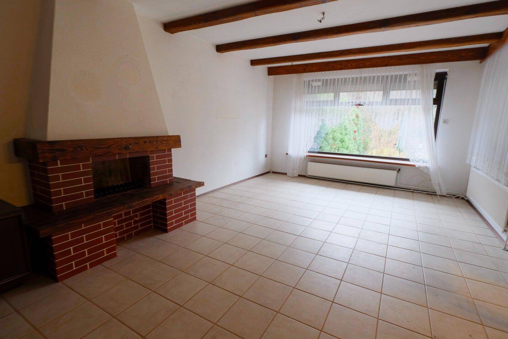 Geräumiges Wohnzimmer mit Kamin, Fliesenboden und großen Fenstern zum Garten.
