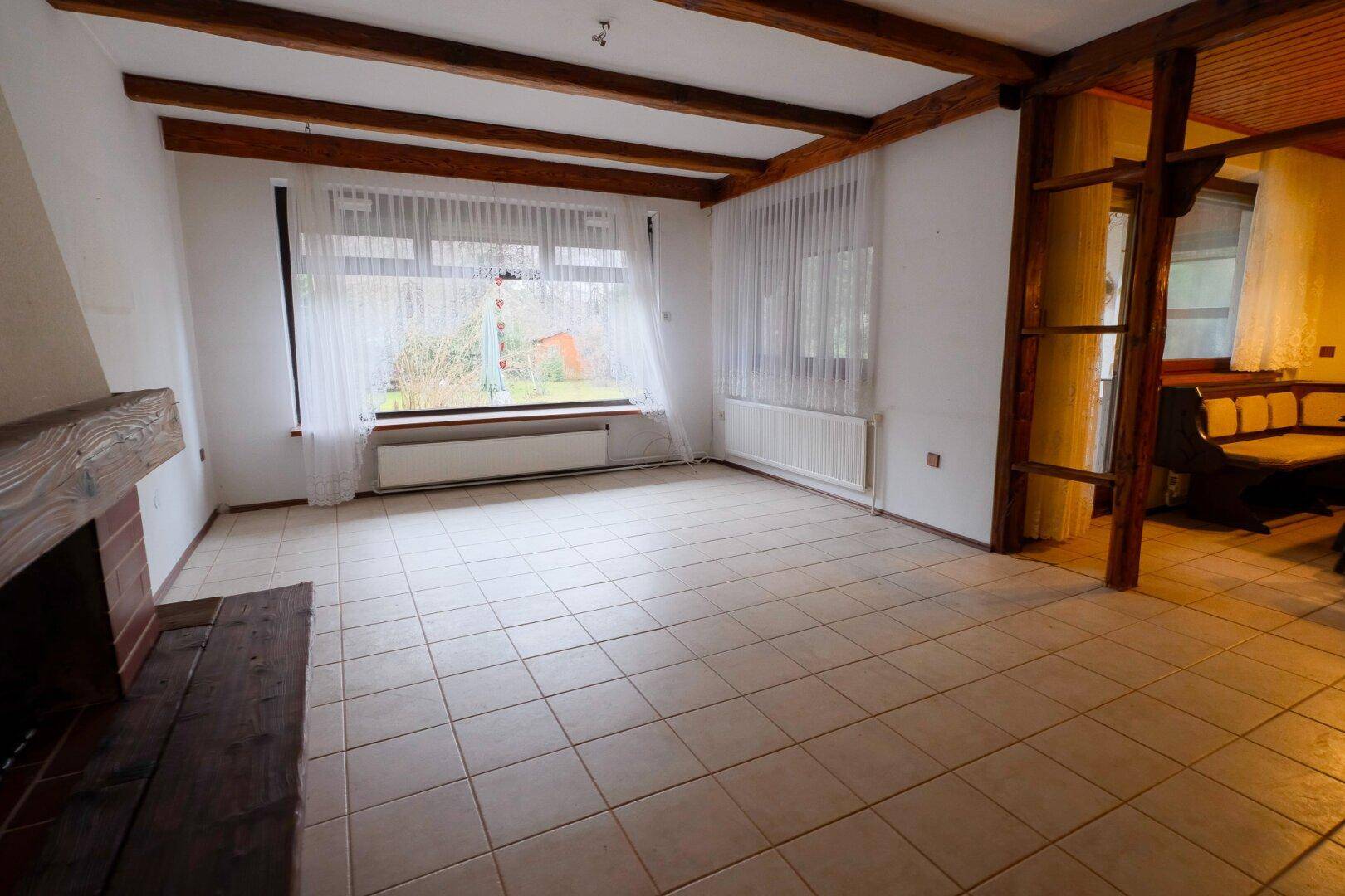Helles Wohnzimmer mit Kamin, Fliesenboden und großen Fenstern mit Blick in den Garten.