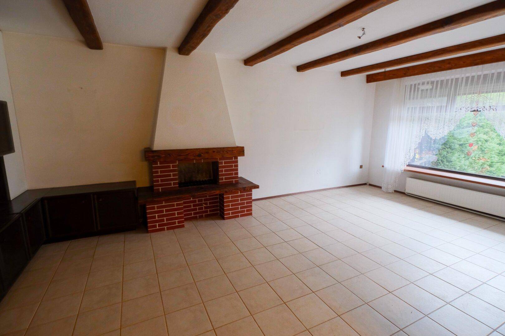 Wohnzimmer mit gemauertem Kamin, Fliesenboden und Holzbalken an der Decke.