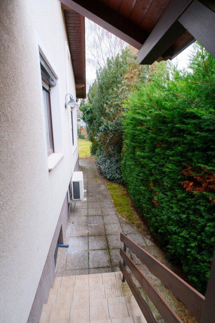 Schmaler Weg entlang des Hauses mit hoher Hecke und Klimaanlage außen.