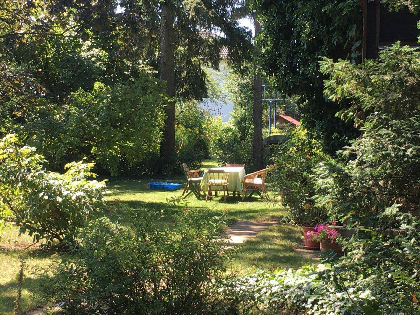 Idyllischer Gartenbereich mit Sitzgelegenheiten unter schattenspendenden Bäumen und blühenden Pflanzen.