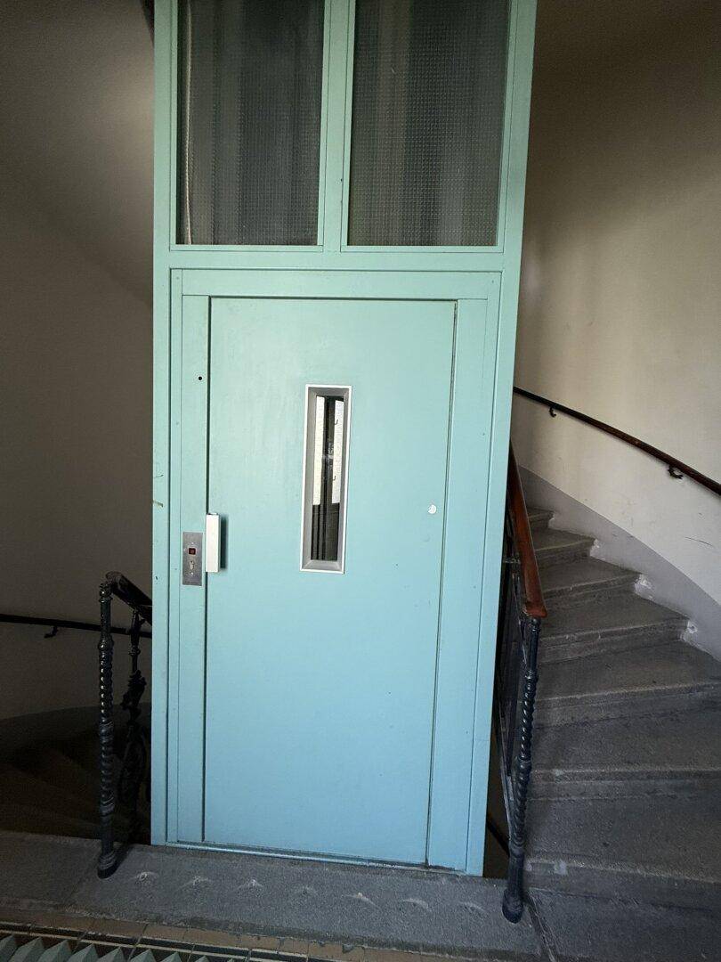 Historischer Aufzug in einem Treppenhaus mit Wendeltreppe, typisch für Altbauten.