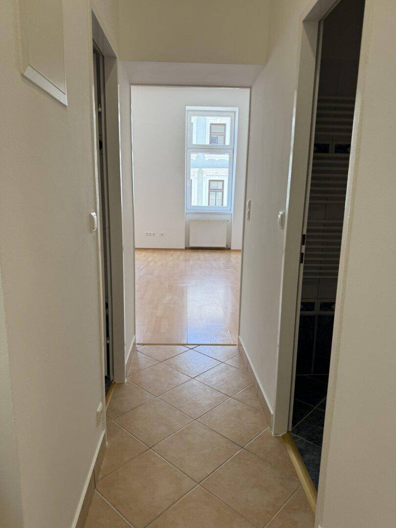 Gefliester Flur, der zu einem hellen Zimmer mit Holzfußboden und Fenstern führt.