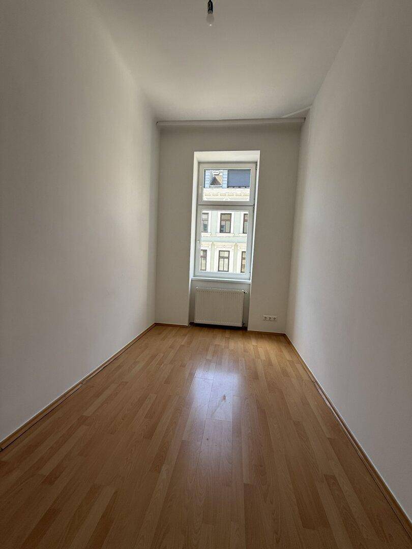 Schmaler, heller Raum mit Holzfußboden und einem Fenster, geeignet als Schlaf- oder Arbeitszimmer.