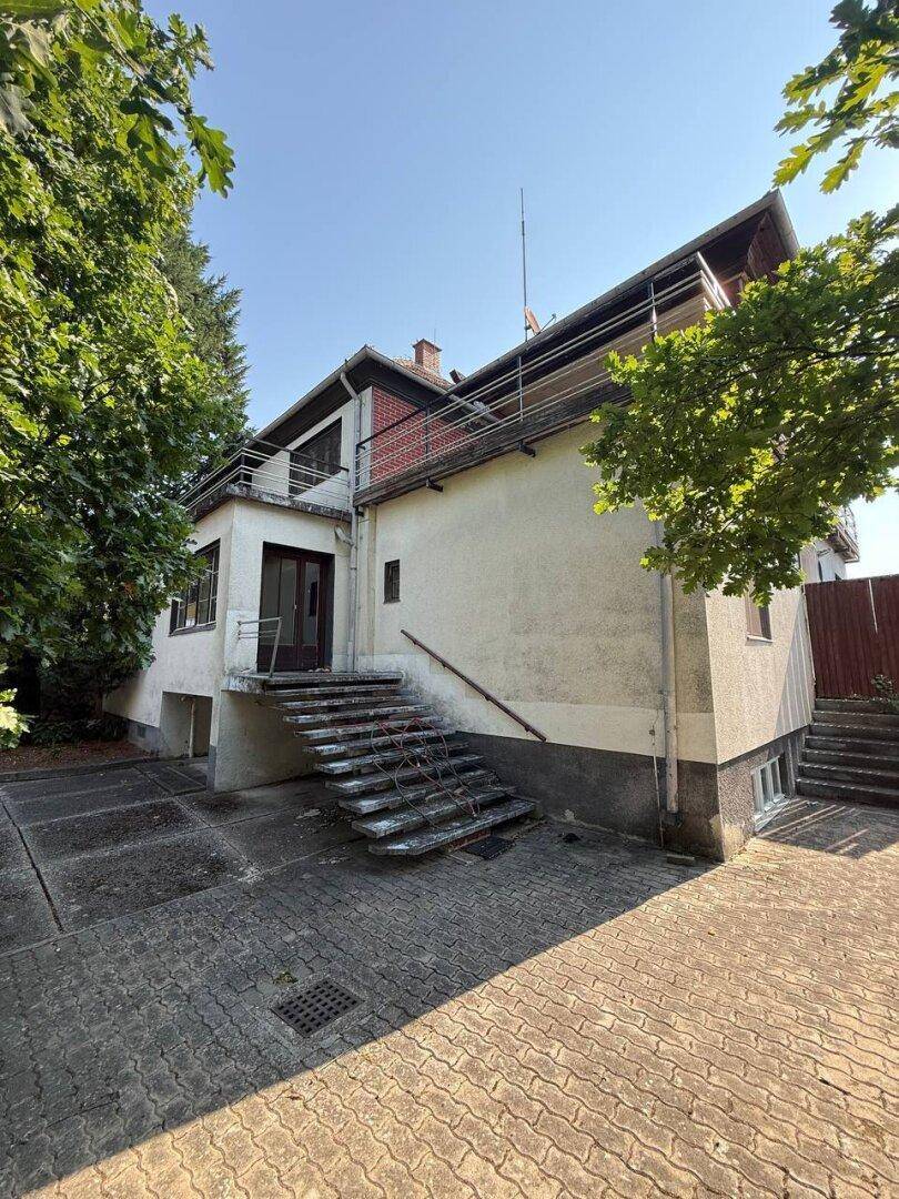Rückseite des Hauses mit Balkon und Außentreppe, umgeben von Bäumen.