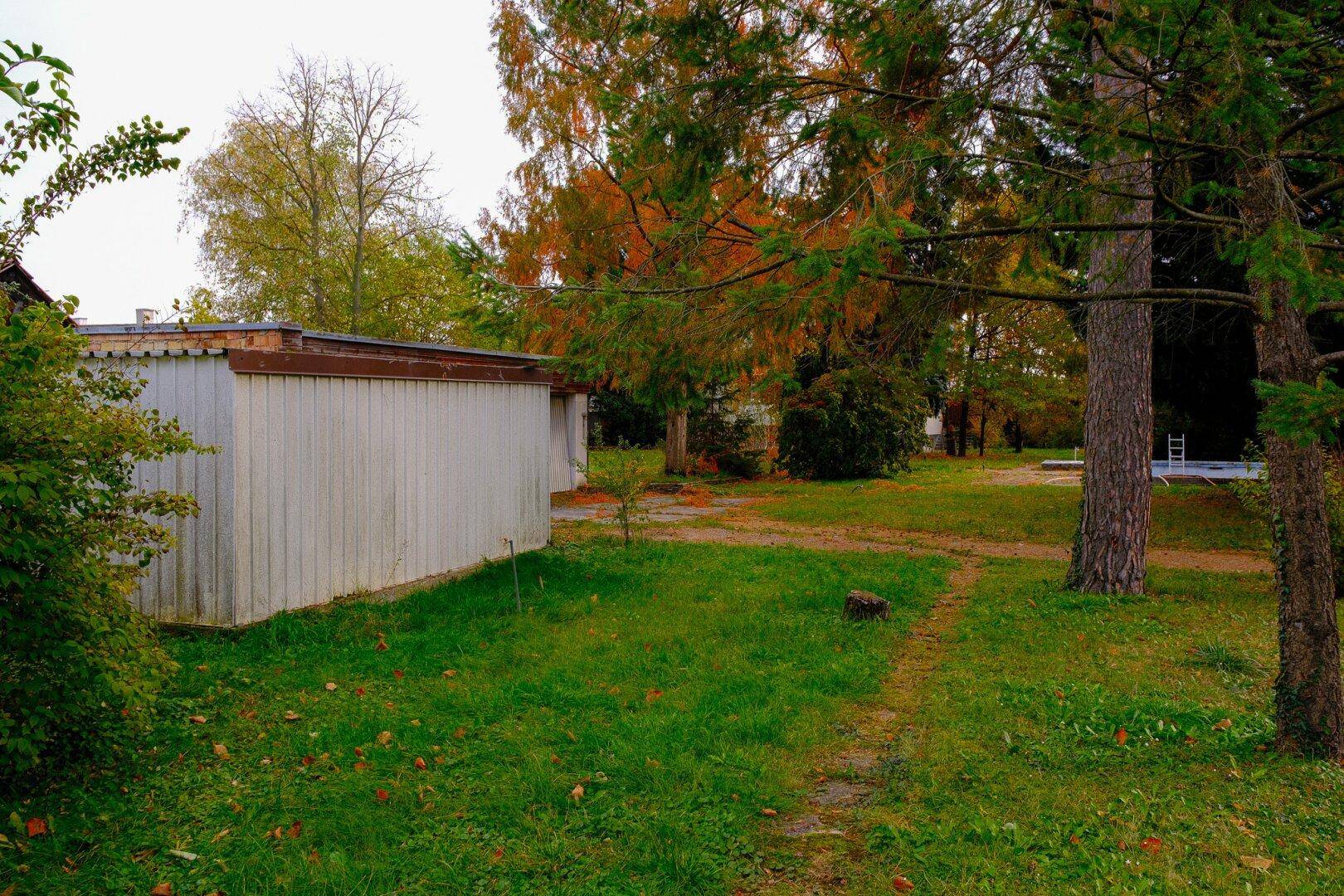 Gartenbereich mit einem weißen Schuppen und hohen Bäumen, bietet viel Grünfläche.