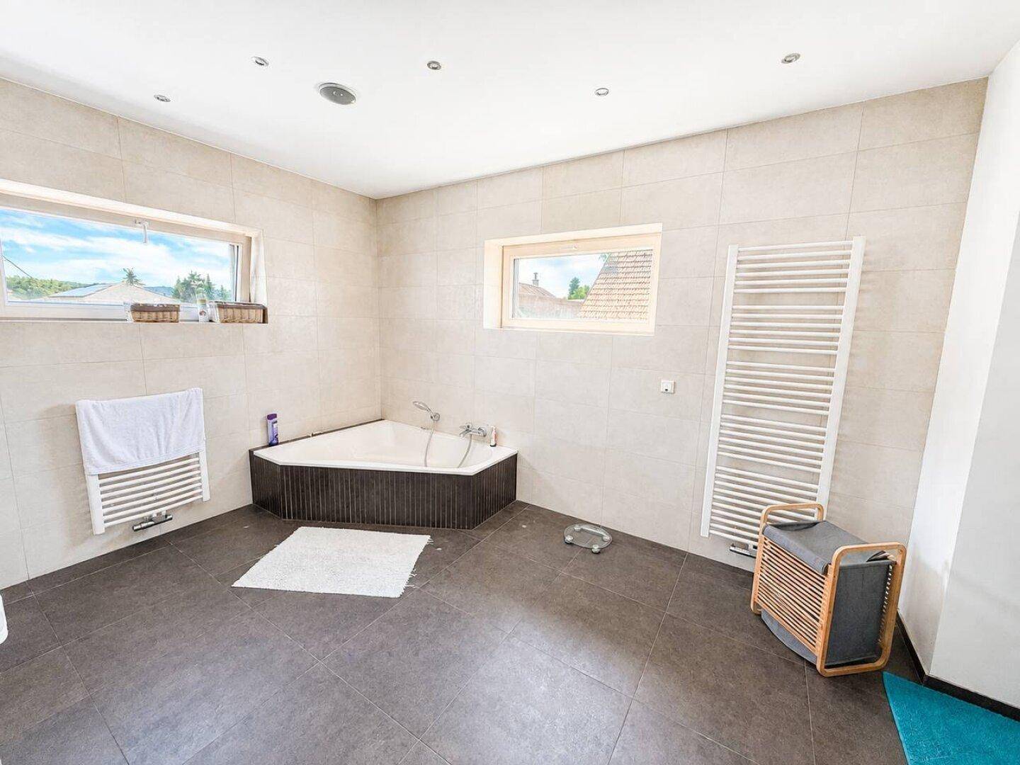 Geräumiges Badezimmer mit Eckbadewanne, beigen Wandfliesen und zwei Fenstern.