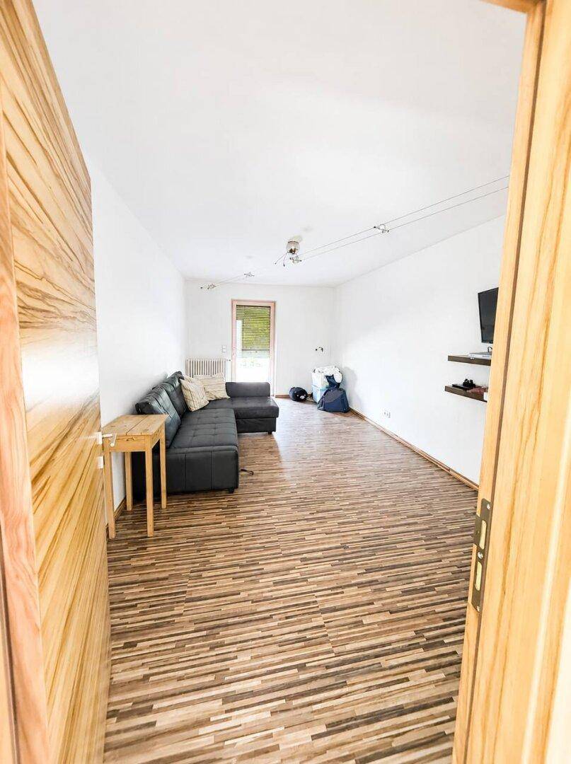 Gemütliches Wohnzimmer mit dunklem Sofa, Laminatboden und Fenster mit Jalousien.