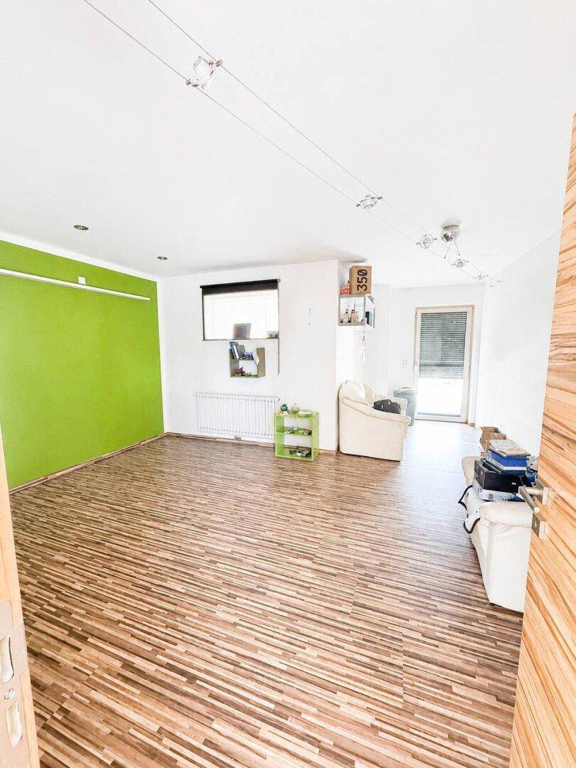 Vielseitiger Raum mit grüner Akzentwand, Laminatboden und Fenster mit Jalousien.