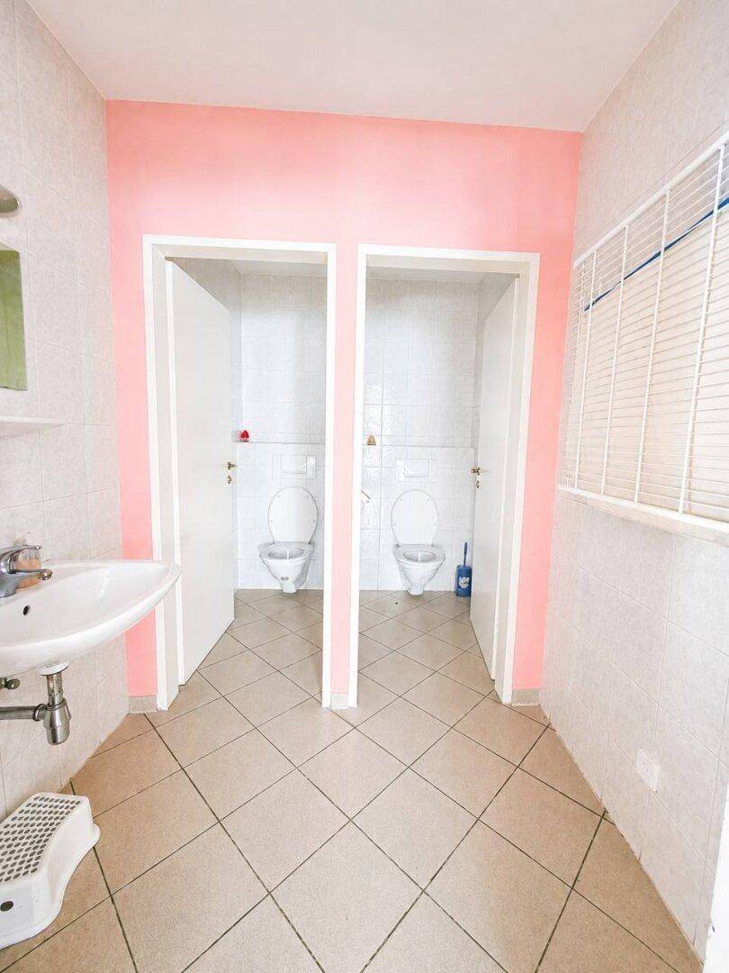 Sanitärbereich mit zwei Toilettenkabinen, einem Waschbecken und rosa gestrichenen Wänden.