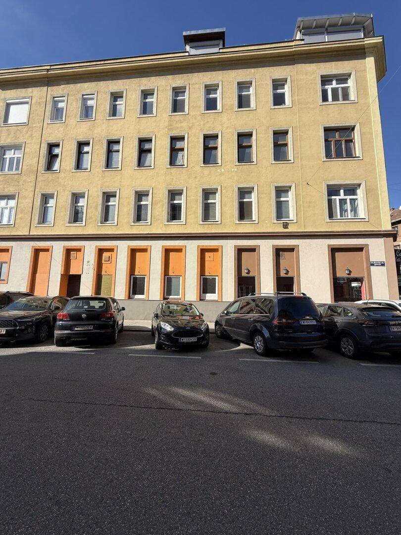 Fassade eines Mehrfamilienhauses mit vielen Fenstern und parkenden Autos auf der Straße.