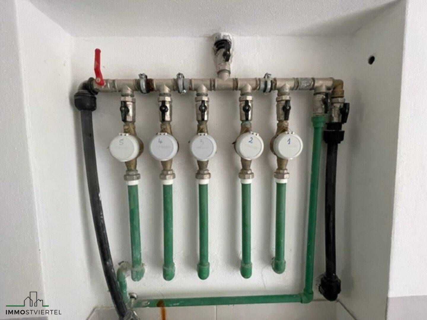 Wasserverteiler mit mehreren Wasserzählern und Rohrleitungen an der Wand.
