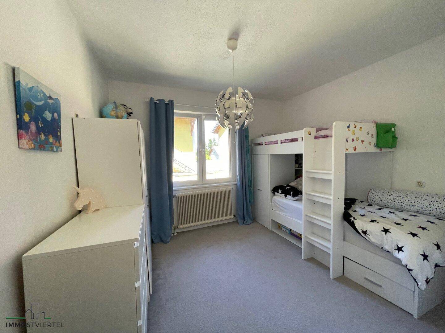 Kinderzimmer mit Etagenbett, weißen Möbeln und einem Fenster mit blauen Vorhängen.