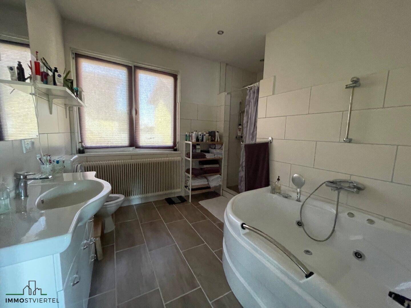 Badezimmer mit Eckbadewanne, Einzelwaschbecken und Duschbereich, hell gefliest.