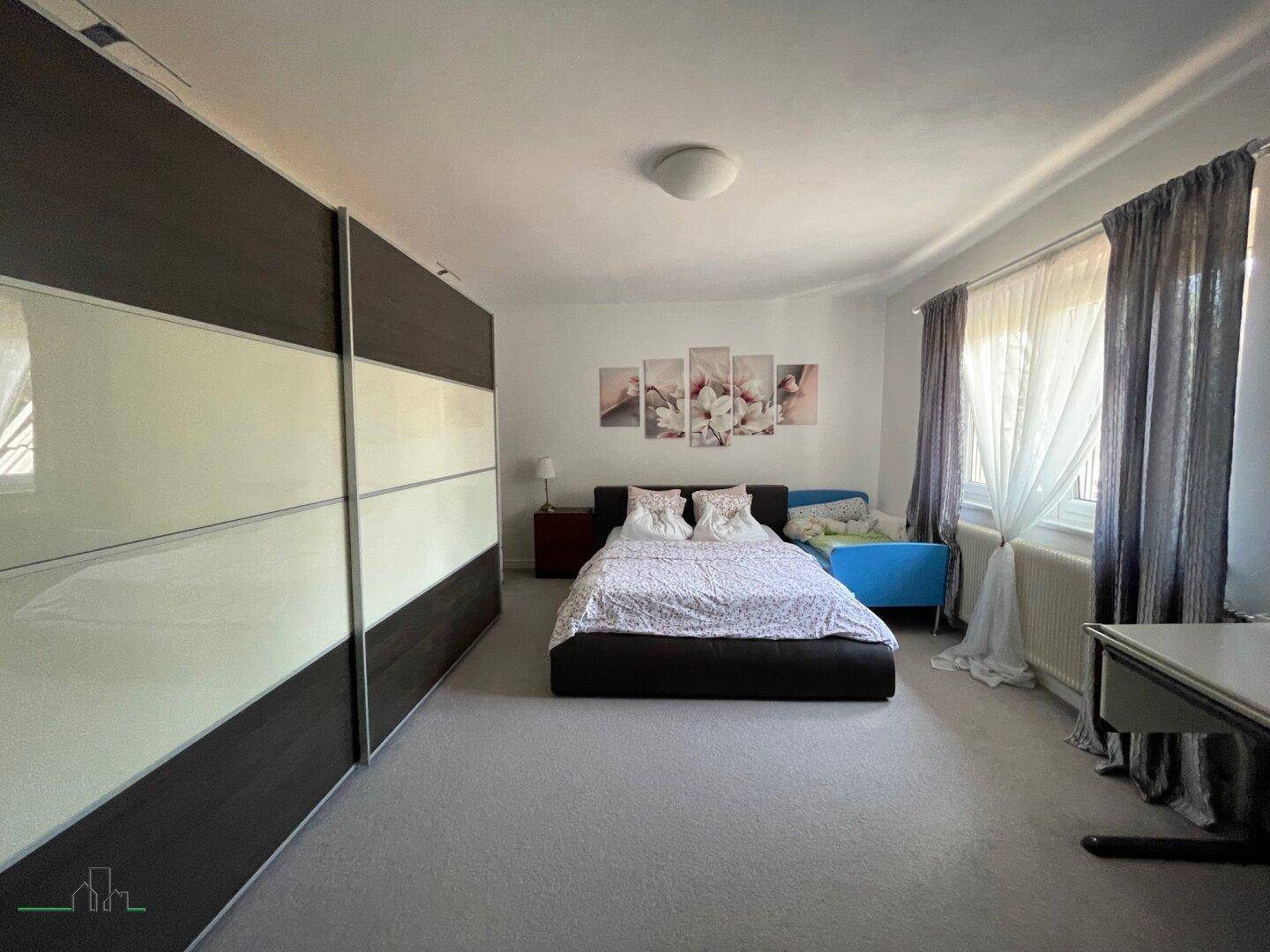 Schlafzimmer mit Doppelbett, großem Schiebetürenschrank und Fenster mit Vorhängen.