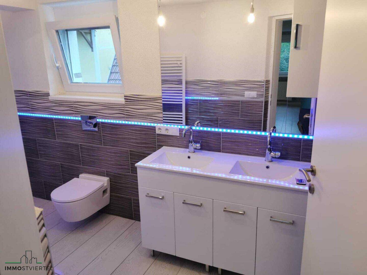 Modernes Badezimmer mit Doppelwaschbecken, Wand-WC, beheiztem Handtuchhalter und Fenster.