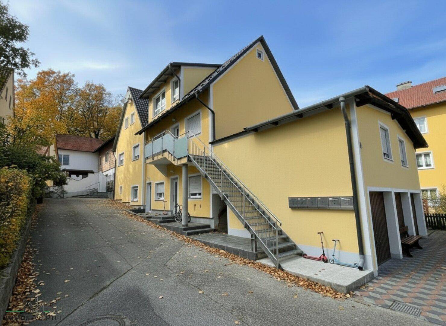Gelbes Mehrfamilienhaus mit externer Treppe und Balkonen, umgeben von herbstlicher Vegetation.