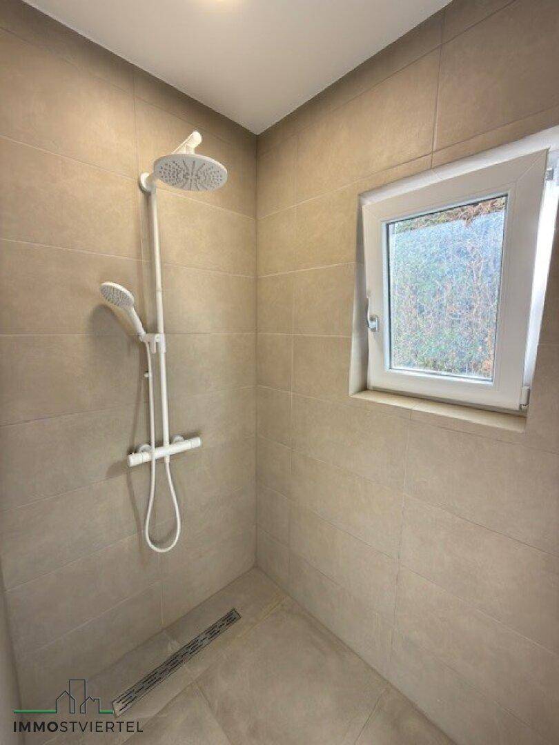 Modernes Badezimmer mit beigefarbenen Fliesen, Regendusche und Fenster für natürliches Licht.