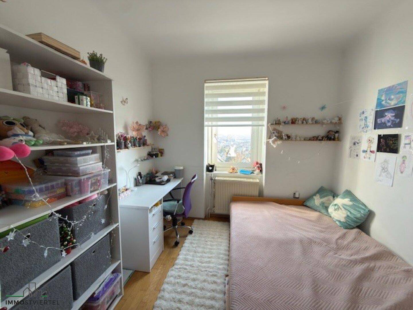 Helles Kinderzimmer mit Schreibtisch, Regal und Bett unter einem Fenster.