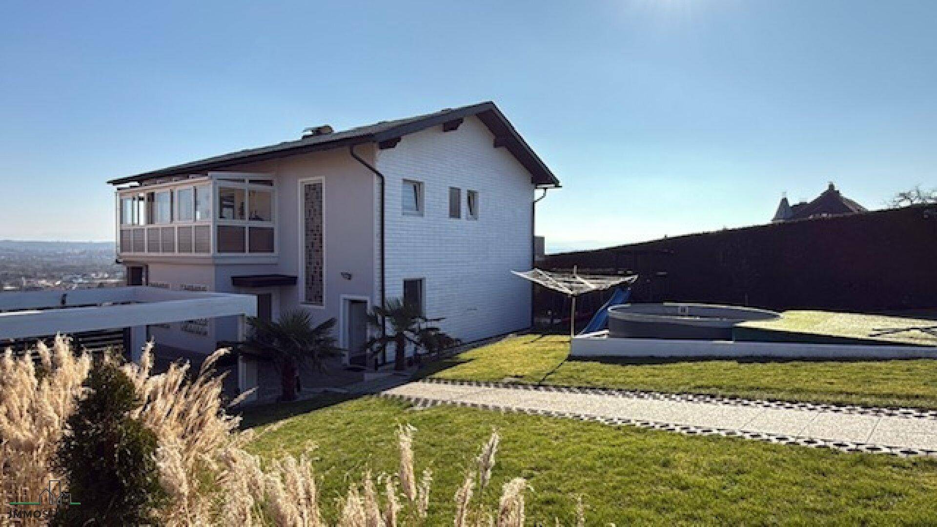 Hausfassade mit Gartenbereich, Pool und Wintergarten unter blauem Himmel.