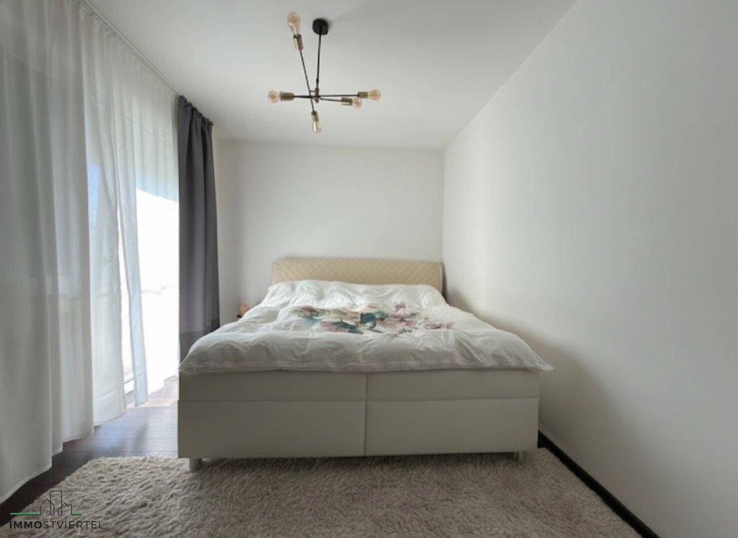 Helles Schlafzimmer mit Doppelbett, Teppichboden und Vorhängen am Fenster.
