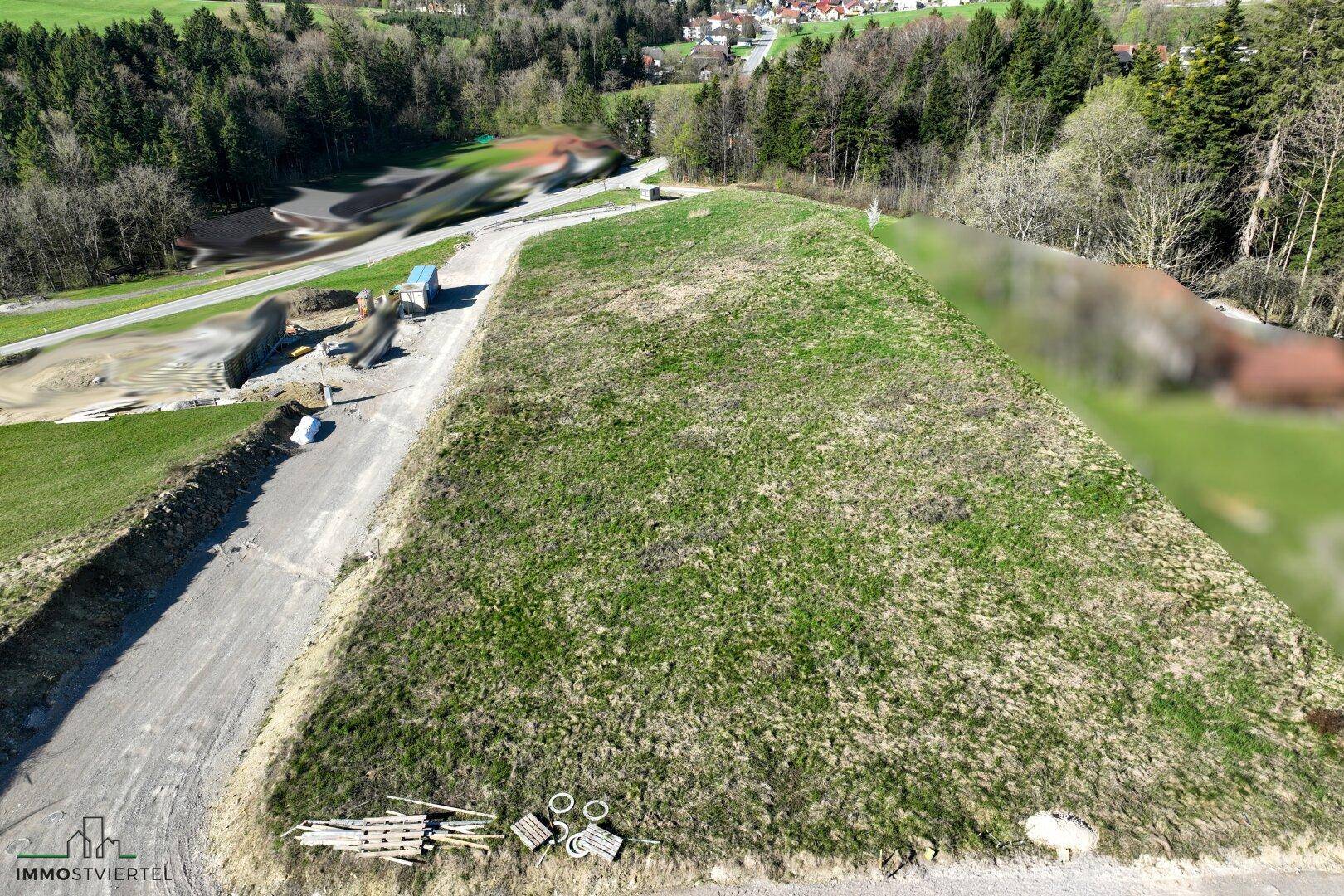 Luftaufnahme eines großen, grünen Grundstücks neben einer Zufahrtsstraße, mit Wald und Häusern in der Ferne.