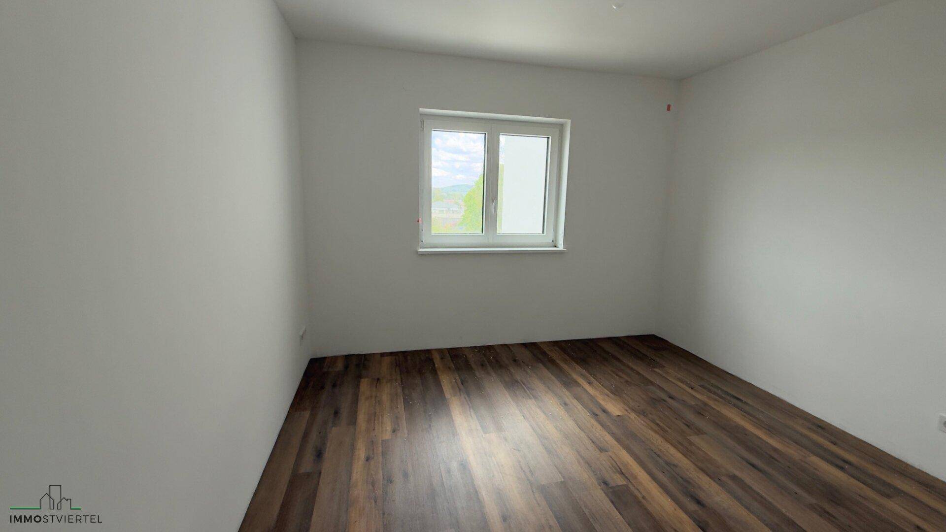 Helles Schlafzimmer mit Laminatboden und einem Fenster mit Blick ins Grüne.