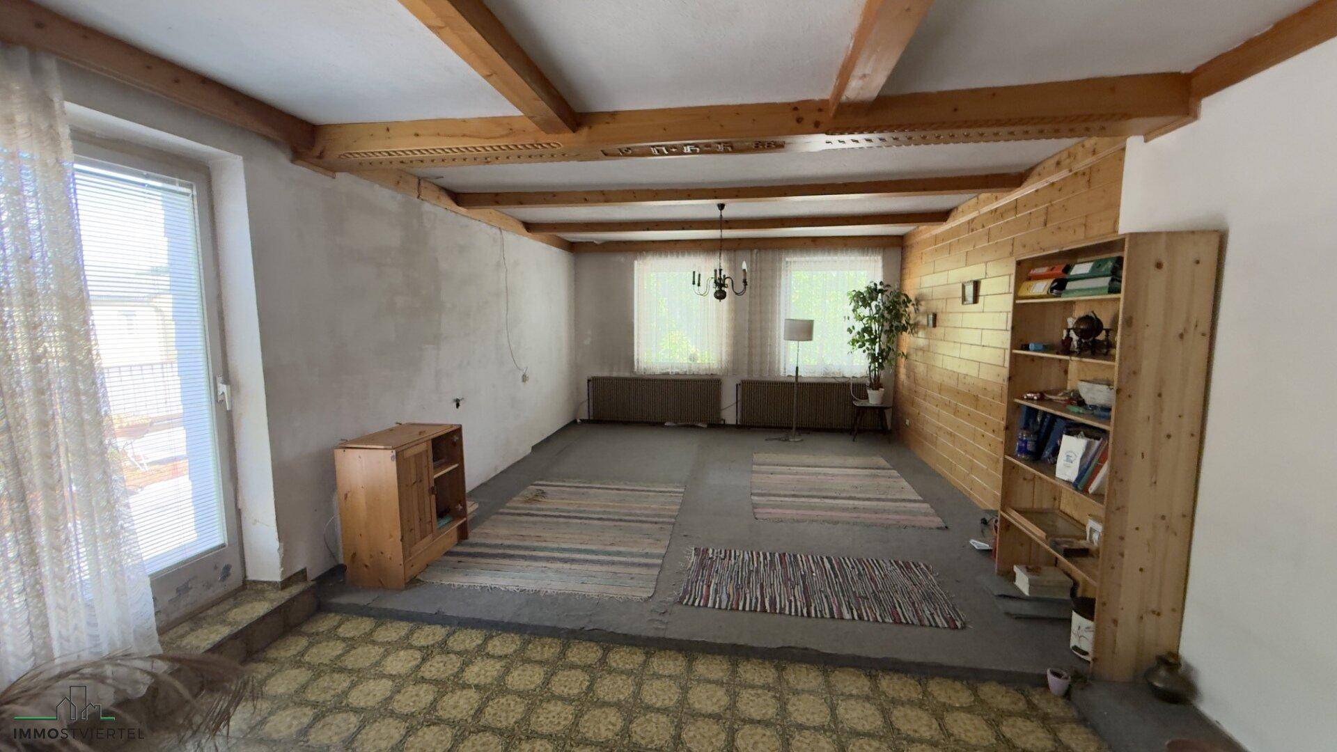 Geräumiges Wohnzimmer mit sichtbaren Holzbalken und großen Fenstern, bereit für die Renovierung.