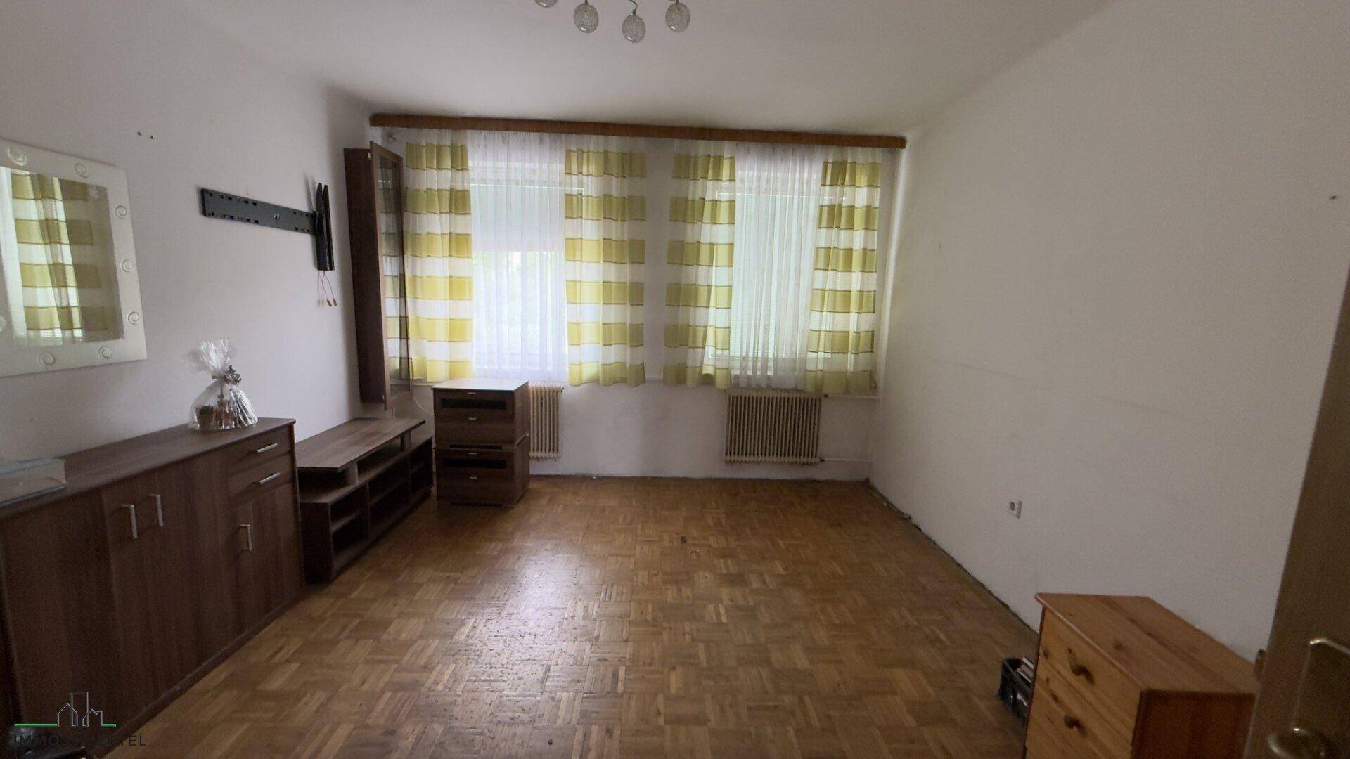 Wohnzimmer mit Parkettboden, großen Fenstern und einfacher Einrichtung, bietet viel Gestaltungspotenzial.