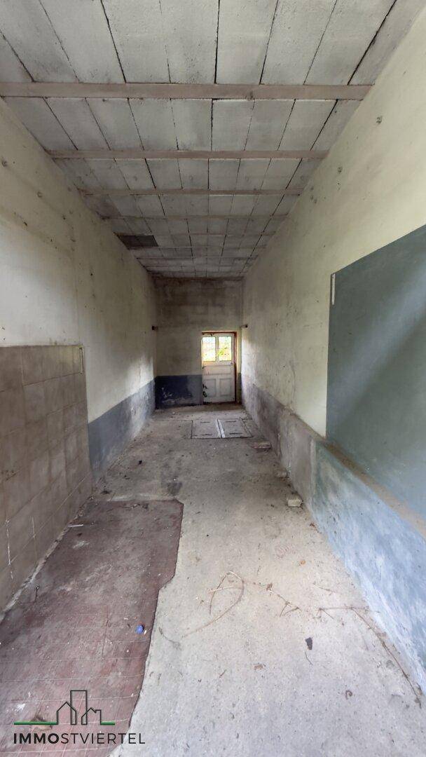 Langer, unfertiger Flur mit Betonboden und sichtbaren Deckenbalken, der zu einem Fenster führt.