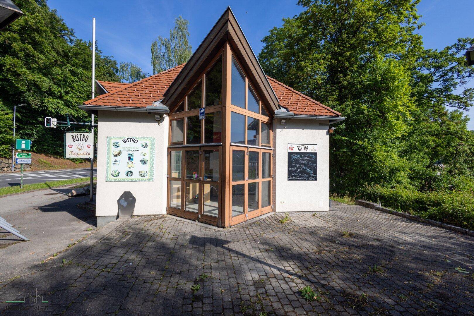 Vorderansicht des Bistros mit großen Glasfenstern, Holzelementen und einem gepflasterten Vorplatz.