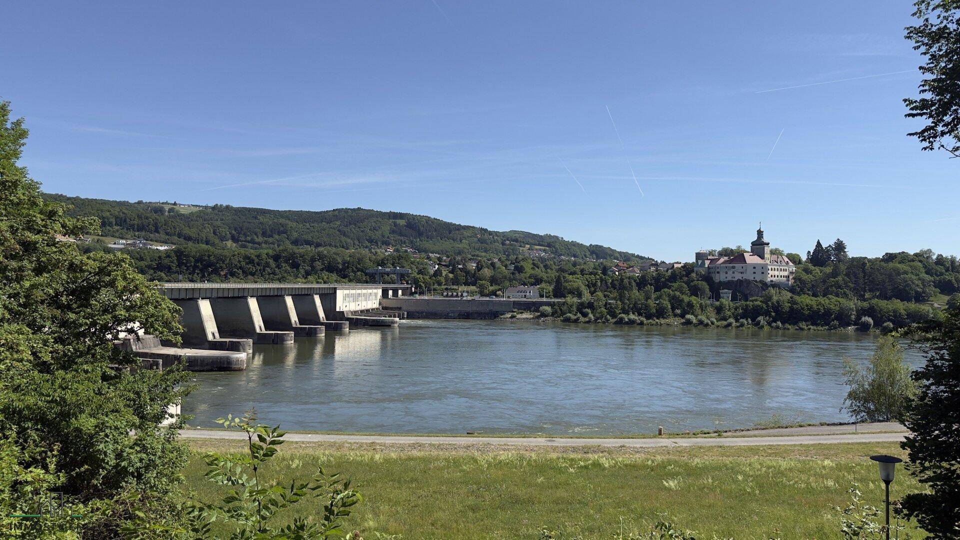 Panoramablick auf den Fluss, das Stauwerk und eine historische Burg am Horizont unter blauem Himmel.