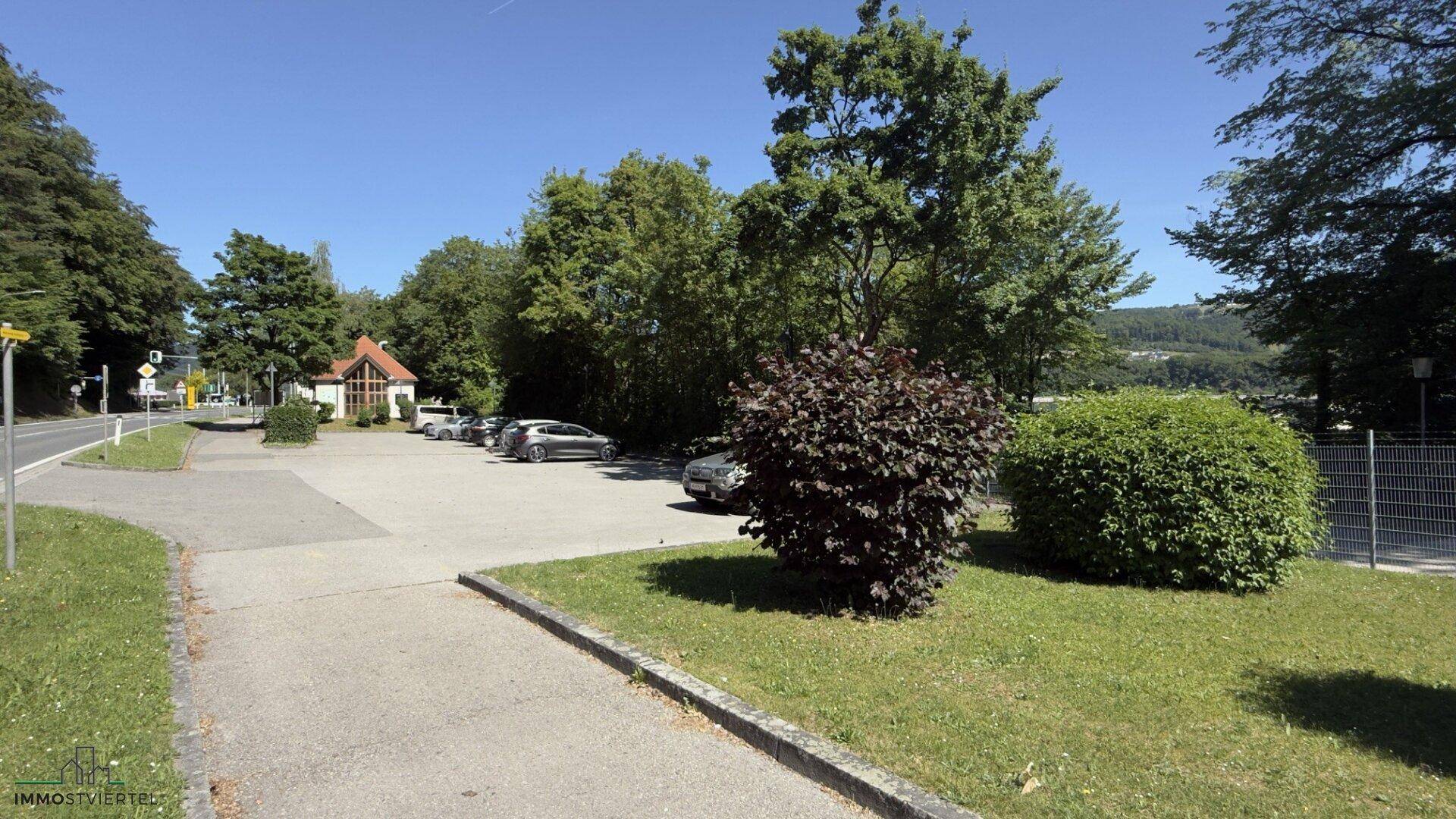Großer Parkplatz vor dem Gebäude, umgeben von Bäumen und Grünflächen an einem sonnigen Tag.