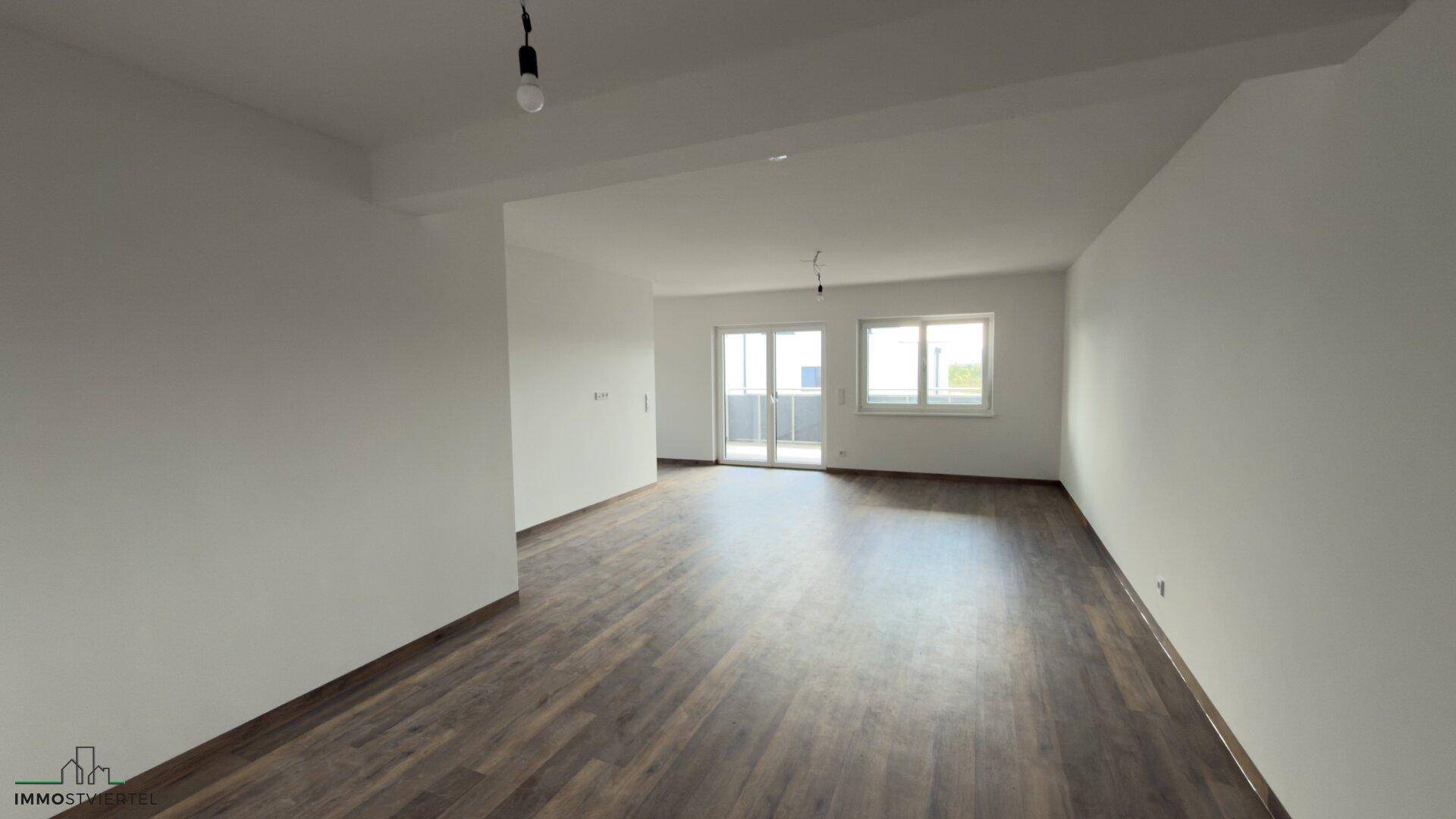 Heller, leerer Wohnbereich mit dunklem Laminatboden und Zugang zu einem Balkon oder einer Terrasse.